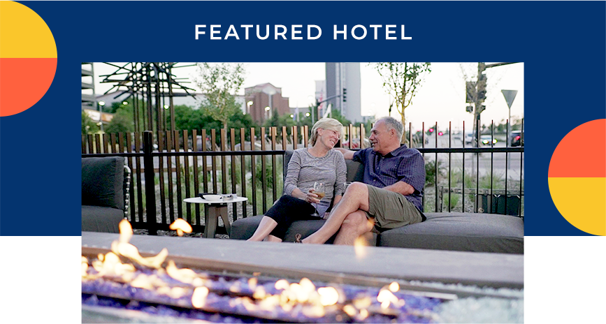 A couple sitting outside on a padded lounge enjoying a glass of wine. A fire feature is in the foreground. A heading reads: Featured Hotel