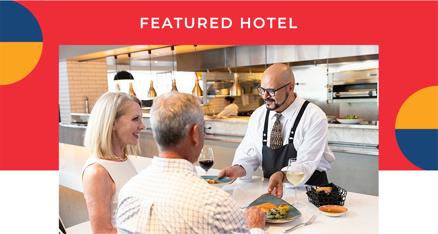 A couple being served elegent looking dishes sitting at the bar. A heading reads: Featured Hotel