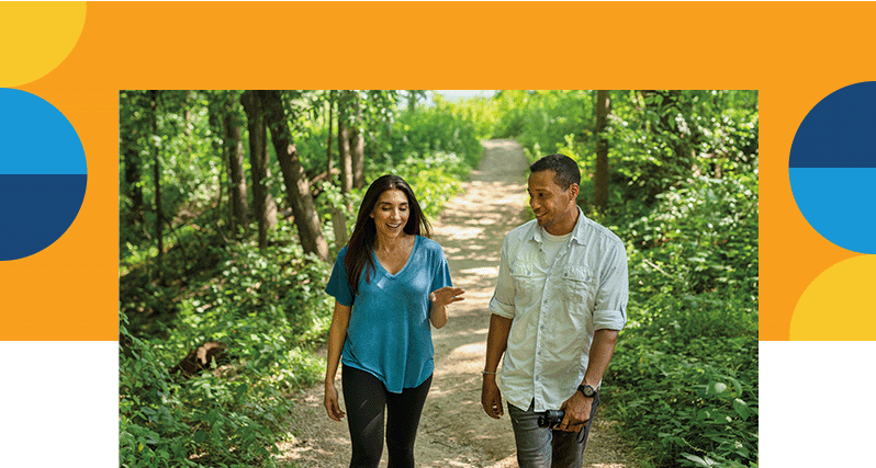 A man and a woman walking on a nature trail surrounded by lush greenery.
